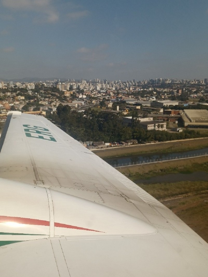 Foto da asa do avião voando, com a pista embaixo e uma cidade distante.