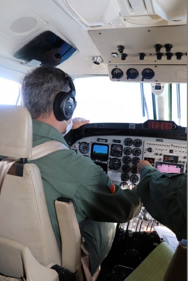 Foto interna do avião, tirada do banco de trás, com o piloto de costas e o painel da aeronave a vista.