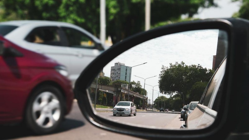Foto fechada de um retrovisor de carro preto, no qual o espelho mostra outra veículo branco vindo logo atrás na rua asfaltada. Ao fundo, em desfoque, passam outros dois carros, um branco e um vermelho.