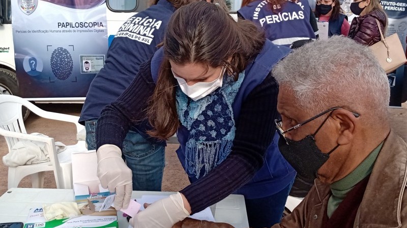 Servidora do IGP, usando luvas de latex, de pé, coletando amostras de sangue do dedo de um senhor que está sentado em cadeira de plástico branco, em frente a uma mesa também de plástico branco, onde aconteceu o mutirão de coletas.