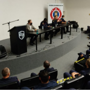 Sala de conferência, com cadeiras enfileiradas e em sua frente, pequeno palco com uma mesa e seis autoridades do evento sentadas, dialogando pelo microfone. No canto esquerdo, bandeiras do Estado, Brasil e Bombeiros.