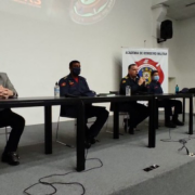 Quatro autoridades oficiais, sentados na mesa disposta no palco da sala de conferência onde ocorre a aula inaugural dos cursos.