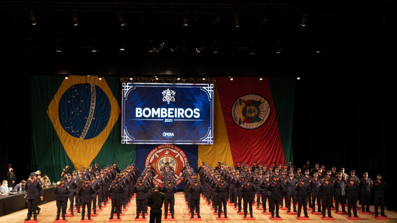 painel identificando os bombeiros, os 101 soldados em formação no palco, fundo com as bandeiras do Brasil, do Rio Grande do Sul e do CBMRS