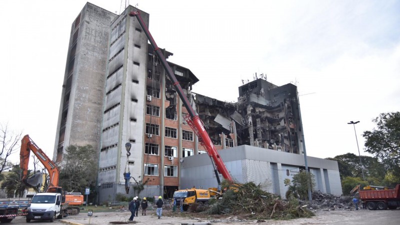 Foto da estrutura colapsada que restou do prédio da SSP após o incêndio de 14 de julho de 2021. Na imagem, também aparecem uma retroescavadeira e um guindaste.