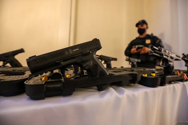 pistolas colocadas em cima  de uma mesa, monitoradas por agentes penitenciários