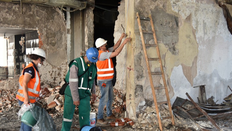 Dois operários com uniformes laranja e capacetes brancos, e um com roupa verde e capacete azul, estão em pé um espaço com paredes de concreto. Um dos operários de laranja segura com as duas mãos fios próximos a um buraco feito um pilar. À direita, uma escada escorada na parede desse pilar.