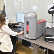 Diante de uma mesa com uma tela de computador e uma grande máquina, o indexador balístico, uma perita trabalha. 