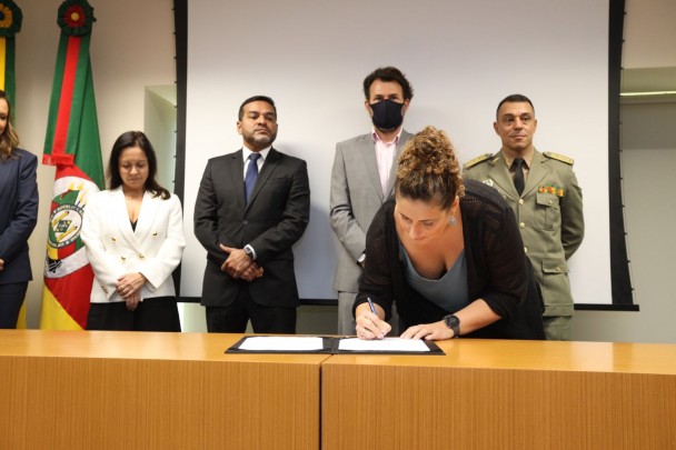 A coordenadora de Parcerias do Instituto Avon, Renata Rodovalho, assina um papel sobre um pasta preta que está em cima de uma bancada de madeira. Atrás dela, outras autoridades observam.