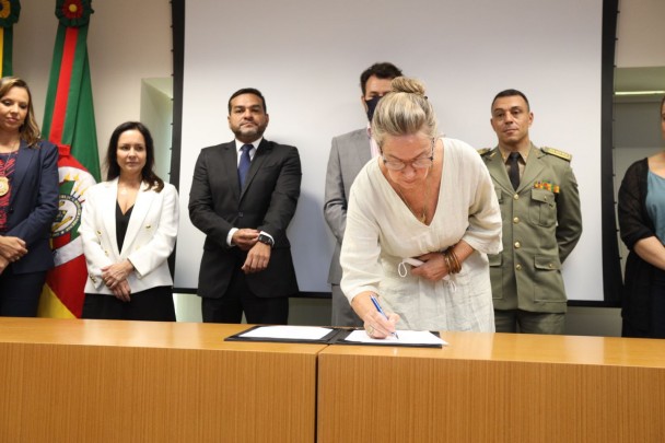 Secretária Regina Becker assina um papel que está sobre uma pasta preta em cima de uma bancada de madeira. Atrás dela, outras autoridades observam.