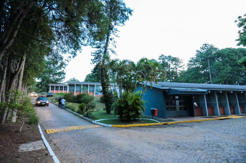 Pátio do complexo da Cidade da Polícia com uma rua lateral à esquerda da foto, toda margeada por árvores, de onde bem ao fundo se veem duas viaturas da Polícia Civil trafegando. À direita da foto, sequência de três edificações, uma atrás da outra do primeiro plano até o fundo da cena.