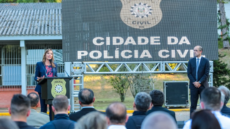 Chefe de Polícia, Nadine Anflor, e subchefe da Polícia, Fábio Motta Lopes, em cima do palco, com um grande telão que mostra o logo da PC e o texto "Cidade da Polícia". Nadine, à esquerda, fala em um púlpito com microfone. Em primeiro plano, desfocada, a plateia que assiste ao discurso.