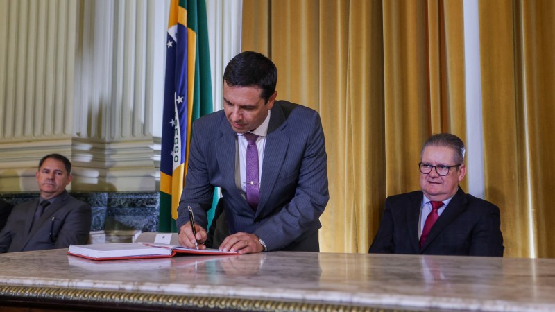 Coronel Vanius Cesar Santarosa, de terno cinza, camisa clara e gravata lilás, assina livro de posse como novo secretário da Segurança, ao lado do governador Ranolfo Vieira Júnior, que está sentado, de terno preto, camisa azul e gravata vermelha. Ao fundo, bandeira do Brasil.