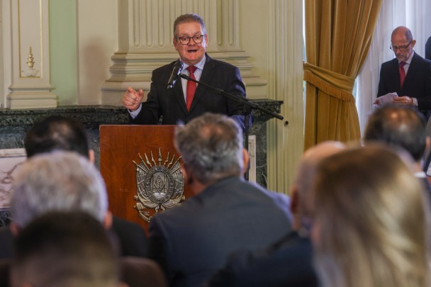 Governador Ranolfo, em pé, de terno preto, camisa clara e gravata vermelha, fala diante de um púlpito com o brasão do estado e um microfone. Em primeiro plano, de costas e fora de foco, pessoas que assistem ao discurso.
