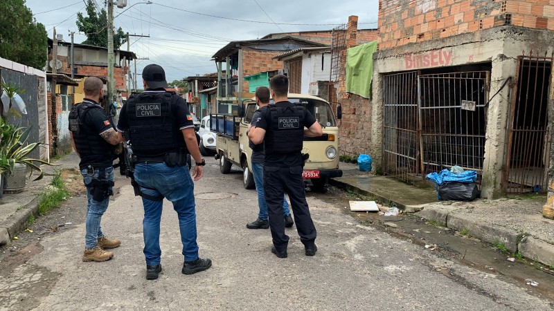 Quatro policiais civis com coletes da instituição aparecem em pé em uma viela de uma vila na zona sul de Porto Alegre. Ao fundo, carros uma kombi e um fusca estacionados.