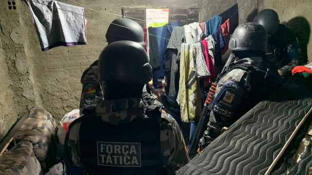 Quatro policiais militares com coletes da força tática e capacetes aparecem em um cômodo de uma casa ao lado de um colchão em pé e próximos de dois varais com roupas penduradas.