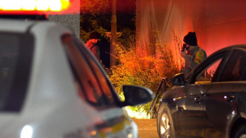 Em primeiro plano, sem foco, parte de uma viatura com giroflex vermelho ligado. Ao fundo, no foco da imagem, um policial militar utiliza um radiocomunicador ao lado de um carro preto.