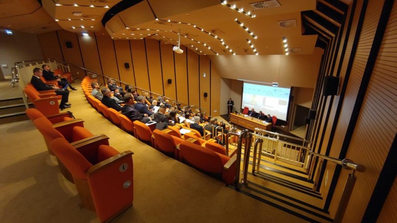 Foto mostra o auditório do TRF4, que é composto por fileiras de cadeiras em formato de arquibancada. A visão é do alto para baixo, onde aparece uma mesa com cinco cadeiras e, atrás, um telão.