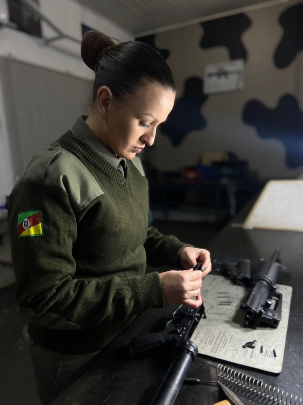A soldado Caroline Chagas Jobim, com cabelo preso em coque e vestindo farda da BM, segura com as duas mãos uma pequena peça de uma arma longa que está desmontada sobre uma bancada a sua frente.