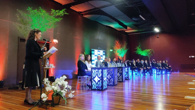 Diretora-geral do IGP, Heloísa Kuser, em pé, fala diante de um púlpito com microfone. Ao lado no palco, a mesa de autoridades da solenidade de formatura e o grupo de formandos sentados lado a lado.