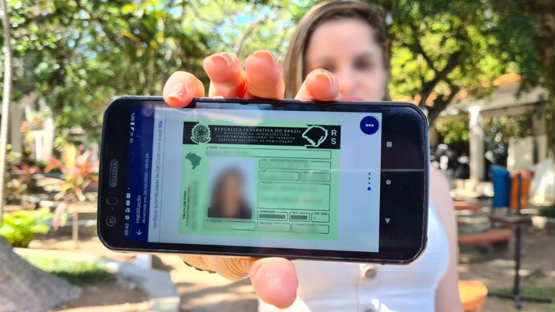 Foto de um celular na horizontal em cuja tela aparece uma CNH digital com os dados de identificação borrados. O aparelho é segura pela mão de uma mulher que aparece desfocada ao fundo em pátio com árvores.