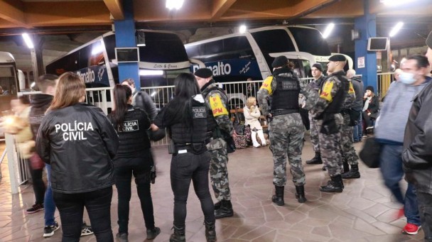 Grupos de policiais civis e militares aparece reunido na plataforma da rodoviária da Capital, onde ao fundo há três ônibus estacionados.