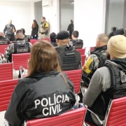 Policias civis e militares aparecem sentados em cadeiras vermelhas dispostas em filas no auditório do Denarc, em Porto Alegre.