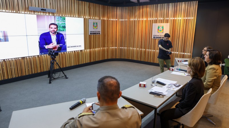 Foto mostra autoridades sentados à mesa enquanto olham para telão com imagem projetada do governador Eduardo Leite durante realização de live.