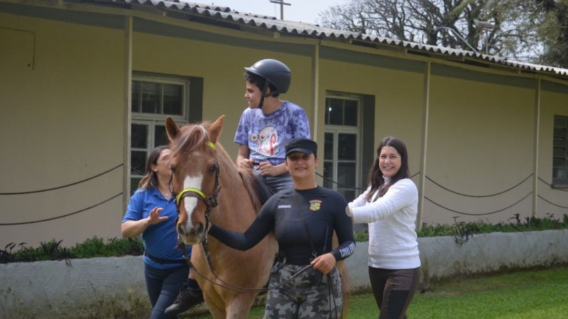 Foto retrata sessão de equoterapia com menino montado em cavalo, acompanhado de três mulheres.