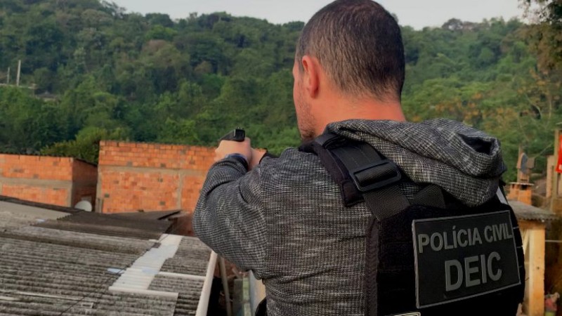 Foto mostra policial de costas com arma em punho sobre telhado de residência