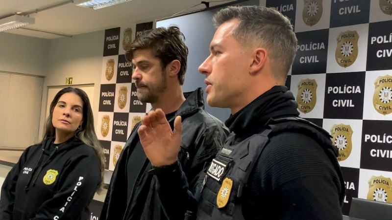 Foto mostra dois homens e uma mulher da Polícia Civil diante de painel da instituição.