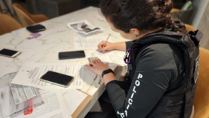 Foto mostra policial mulher escrevendo em papel sobre mesa onde estão outros papéis e aparelhos celulares.