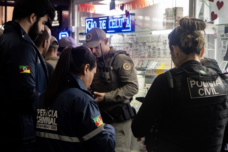 Foto mostra agentes do IGP, da PC e da BM manuseando celulares diante de vitrine de comércio.