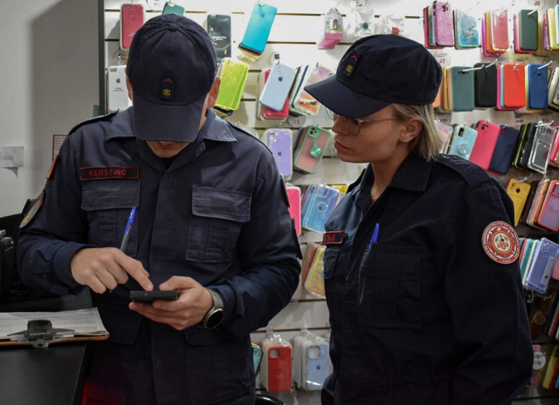 Foto mostra bombeiro e bombeira manuseando celular com vitrine de capinhas de celular ao fundo.