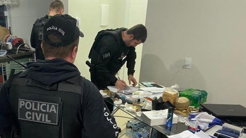 Foto mostra três policiais civis em interior de residência. Ao centro, se vê mesa de vidro com laptop, papéis e outros itens.
