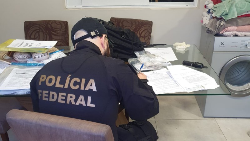 Foto mostra mesa de vidro com papéis e policial federal sentado de costas. Ao fundo, se vê máquina de lavar.