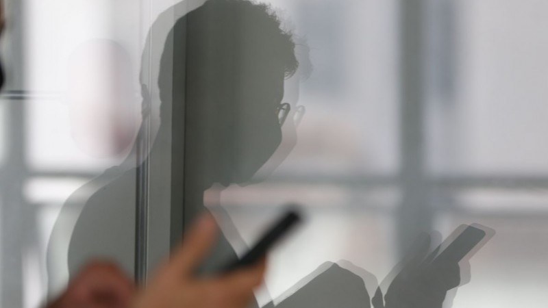 Foto mostra vidro com reflexo de homem olhando para celular. 