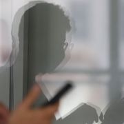 Foto mostra vidro com reflexo de homem olhando para celular. 
