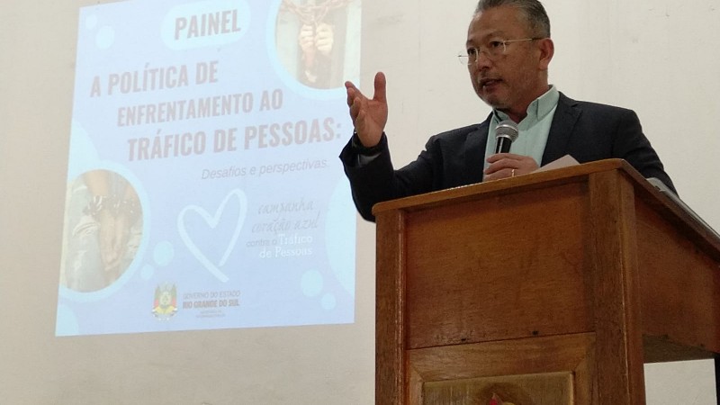 Foto mostra coronel Mário Ikeda com microfone e em pé em púlpito. Ele tem uma das mãos erguidas ao falar com o público. Ao fundo, se vê projeção com nome do painel “A Política Nacional de Enfrentamento ao Tráfico de Pessoas: Desafios e Perspectivas”