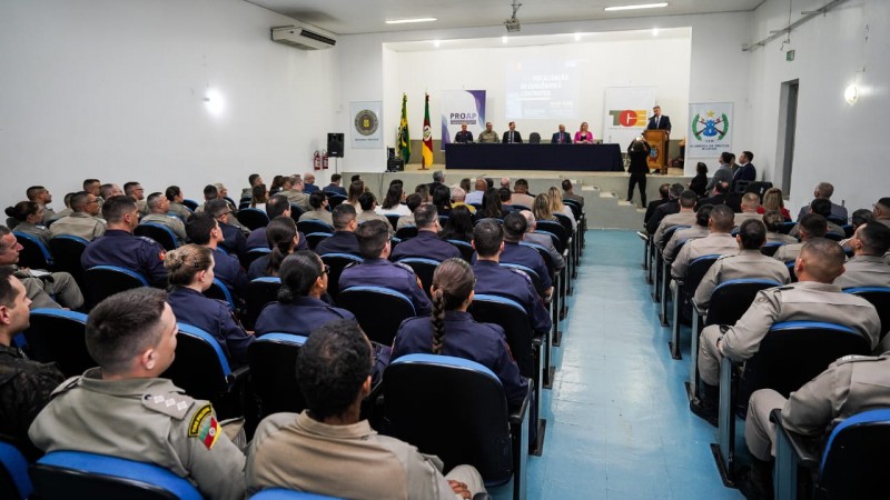 Foto mostra auditório. Público é fotografado de costas, sentado. No palco, se vê autoridades sentadas à mesa.