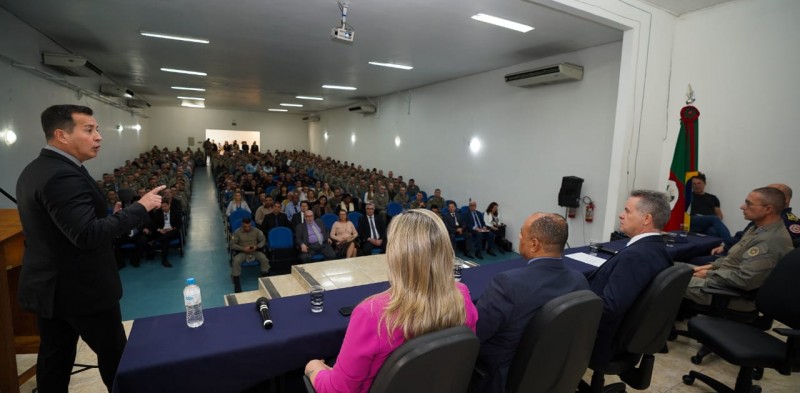 Foto mostra secretário da segurança pública, Sandro Caron, em pé em palco. Outras autoridades estão sentadas à mesa no palco. Ao fundo, se vê público em auditório.
