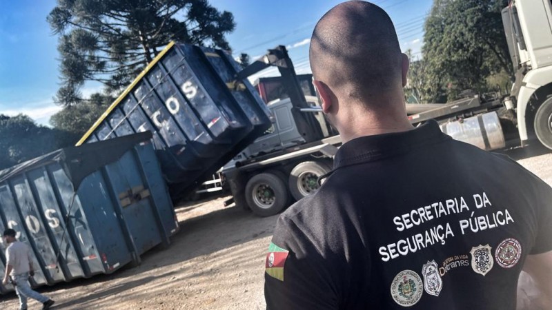 Foto mostra homem de costas com camiseta da Secretaria da Segurança Pública. Ele está em espaço aberto. Ao fundo, se vê parte de caminhão.
