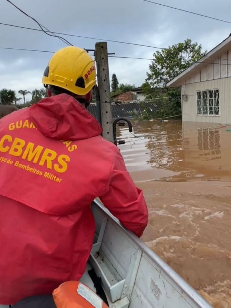 Foto mostra bombeiro de costas, dentro de barco, em rua alagada. Ao fundo, se vê casa.