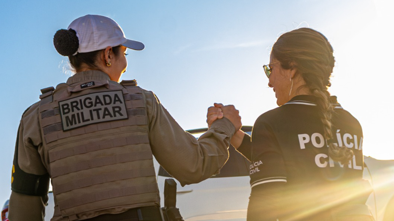 Foto mostra uma policial da BM e outra da PC de costas. Elas se cumprimentam com a mão e olham uma para a outra. Ao fundo, se vê o céu.