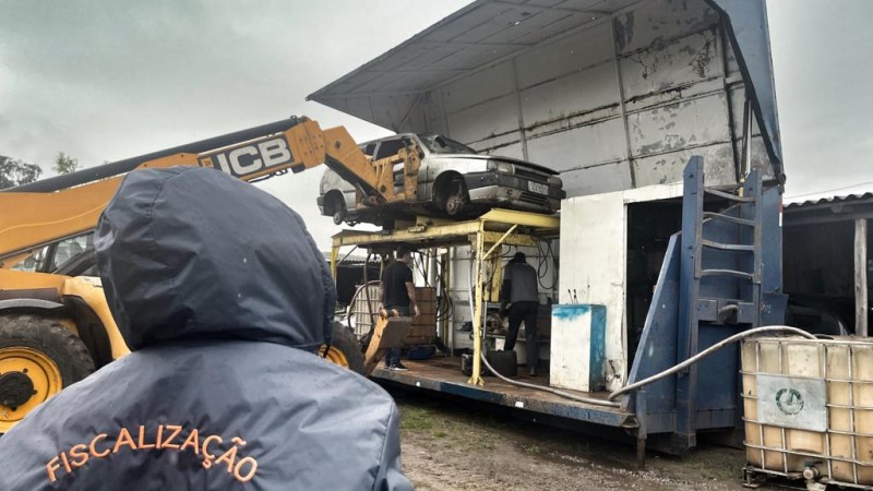 Foto mostra homem com colete do DetranRS diante de carcaça de carro suspensa por máquina.