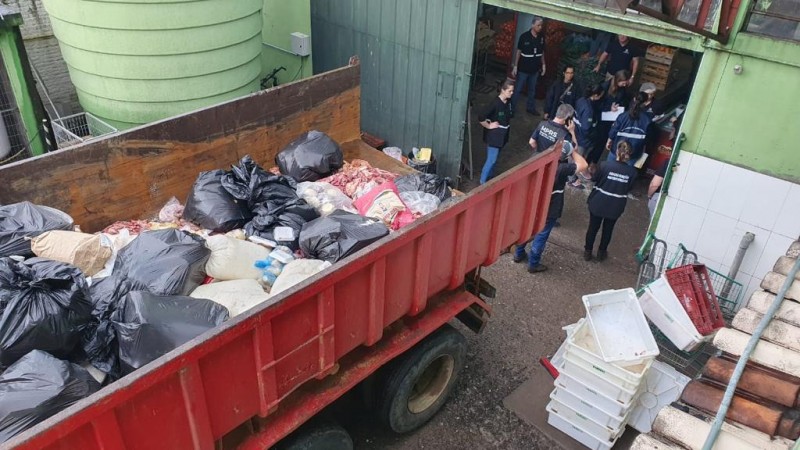 Imagem mostra caçamba vermelha de caminhão cheia de sacolas plásticas com alimentos apreendidos. Ao fundo, se vê grupo de policiais e servidores públicos reunidos.