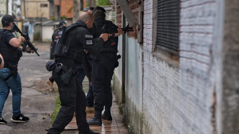 Foto horizontal mostra policiais armados ingressando em moradia de alvenaria.