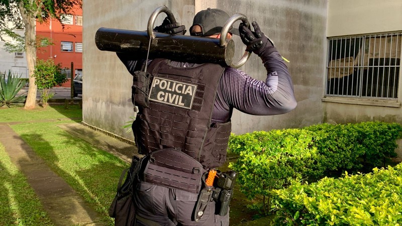 imagem mostra um policial civil equipado com uma farda da policia, colete a prova de balas e uma barra de ferro nas costas