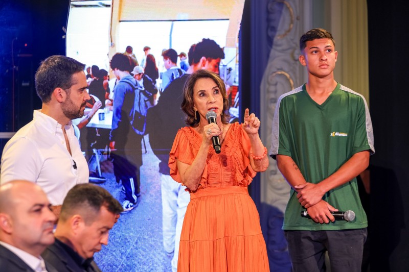 Leite, mais à esquerda da imagem, ouve atentamente a secretária da educação, que está falando ao microfone. Ao lado dela, mais à direita está um dos alunos da rede estadual, que contou sua experiência como estudante da rede. Atrás deles, há uma tela vertical com imagens do ambiente escolar.