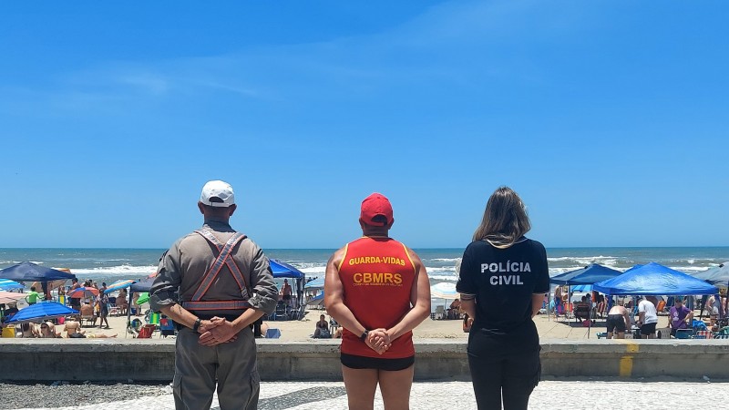 Foto mostra policial militar, guarda-vidas e policial civil lado a lado. Estão em praia. Estão de costas para o fotógrafo e olham em direção ao mar. Na areia, há barracas e banhistas.
