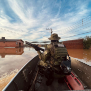 Foto mostra policial militar de costas, empunhando arma. Ele está sentando em barco que navega por área alagada de Novo Hamburgo. A água é lamacenta.
